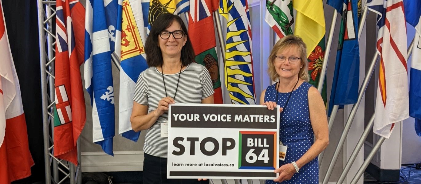 LRSD Board Vice-chair Sandy Nemeth and LSSD Board Chair Lena Kublick holding a Stop Bill 64 sign in front of provincial flags.